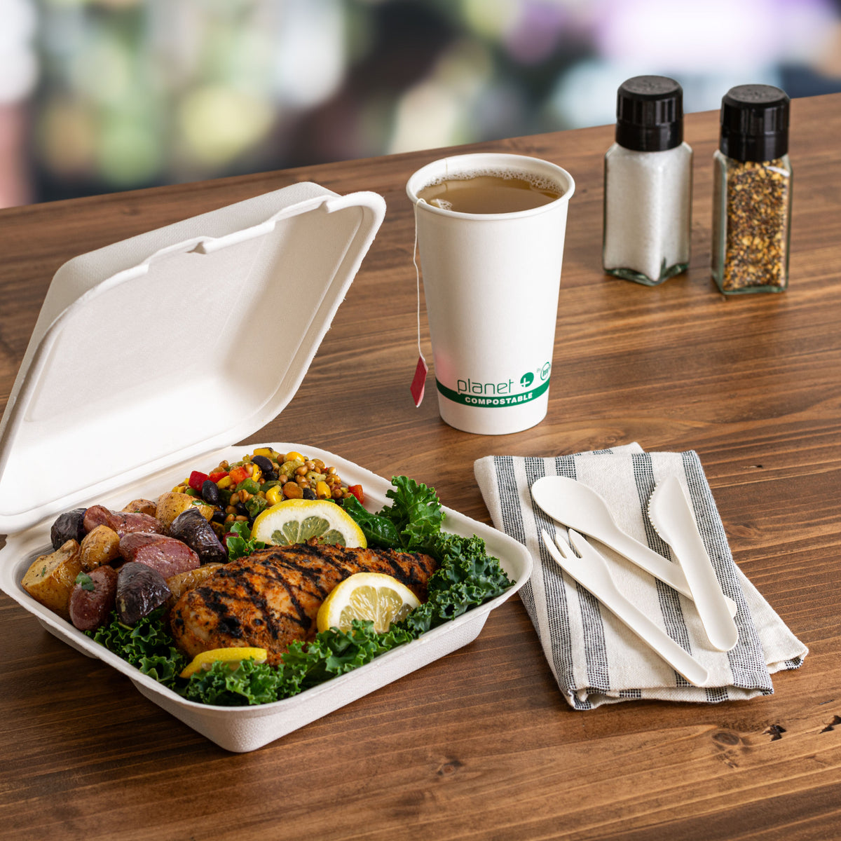 Compostable Paper Cutlery Set on a napking with a compostable clamshell of food and a compostable cup of tea on a wood surface.
