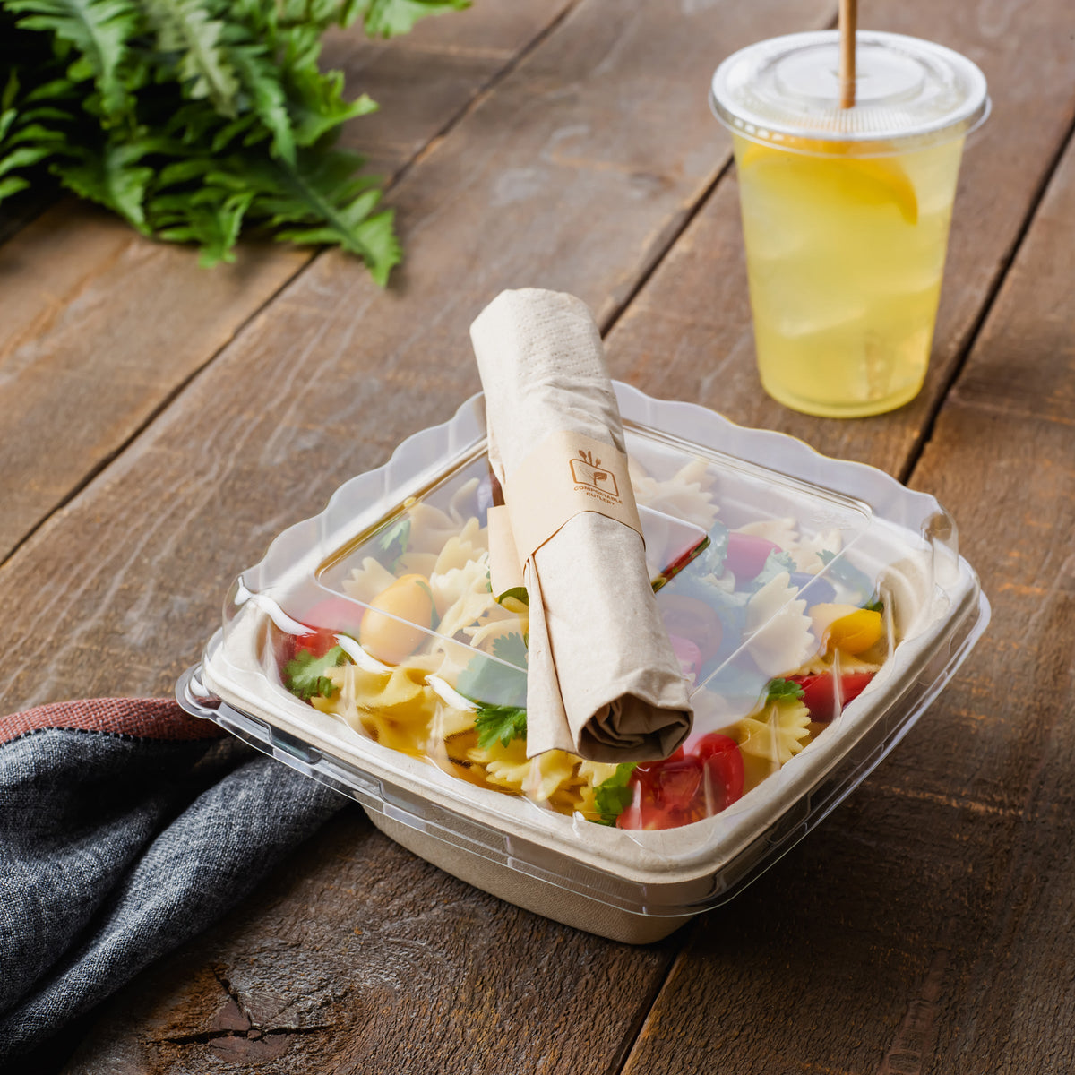 Clear plastic container with rolled compostable cutlery on a wooden table next to a glass of lemonade.
