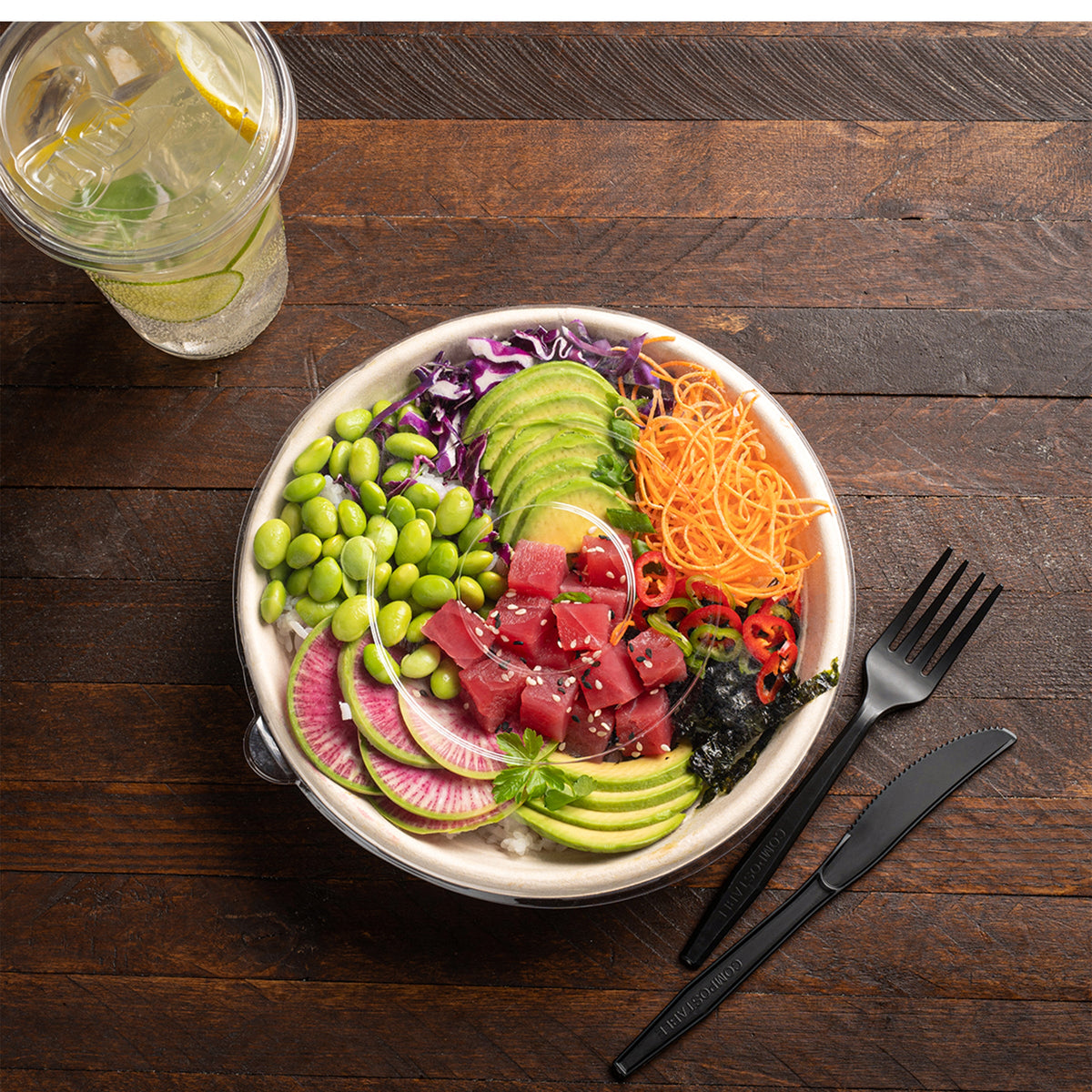 Colorful salad with various ingredients in a disposable bowl on a wooden table with a glass of lemonade.