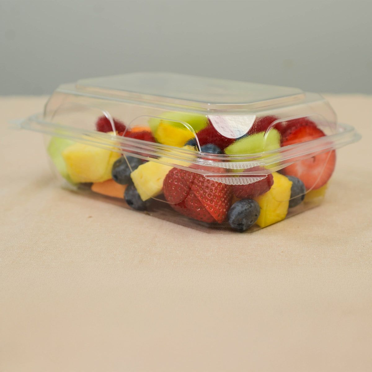 Clear hinged container of mixed fruits on a beige surface with a gray background