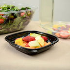 Black bowl of fruit salad on a table with a salad and berries in the background.