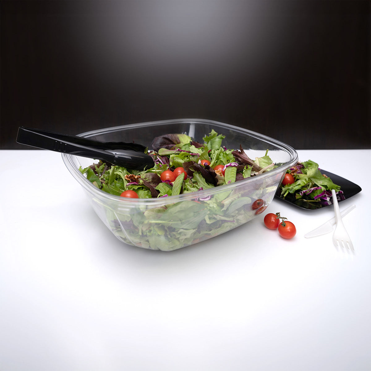 Clear plastic container with salad and black utensils on a white surface