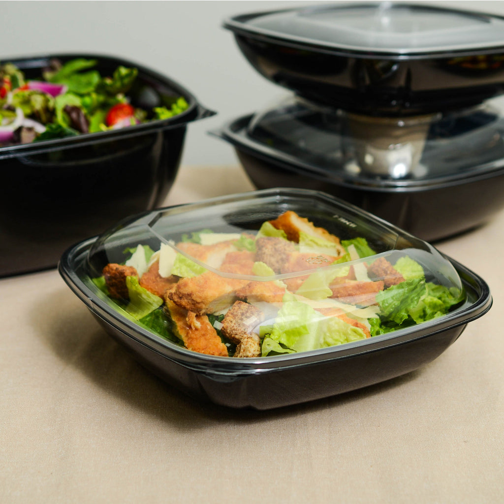 Black plastic containers with clear lids containing salads on a beige surface