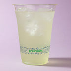Clear compostable cup with greenprint branding filled with a yellowish drink on a light purple background