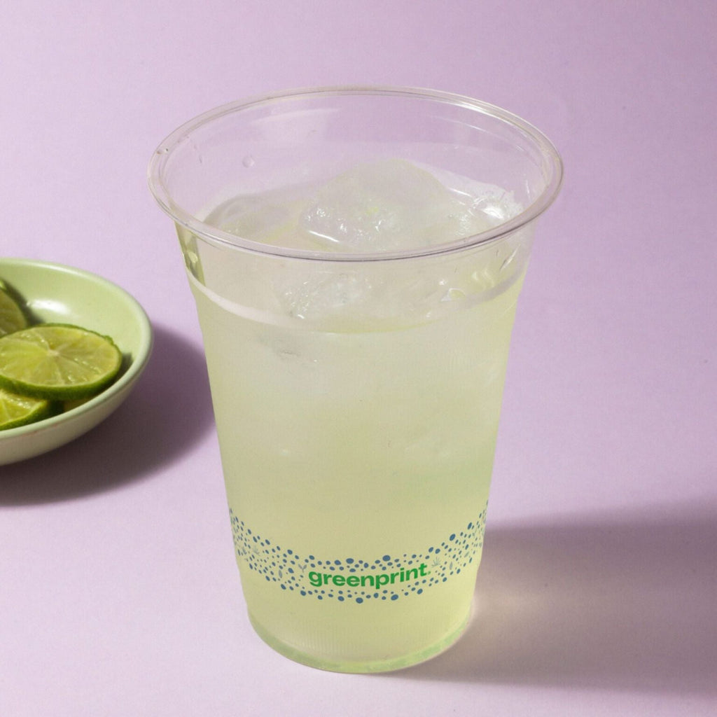 Clear plastic cup with green drink and 'Greenprint' branding, next to a bowl of lime slices on a light purple background.
