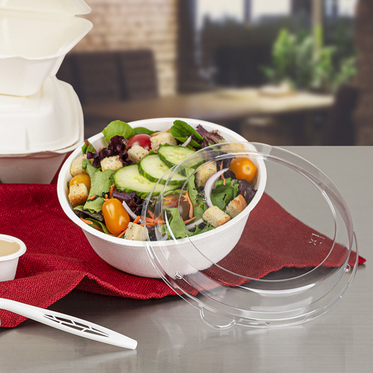 Disposable bowl of salad with a clear lid on a red cloth, blurred background