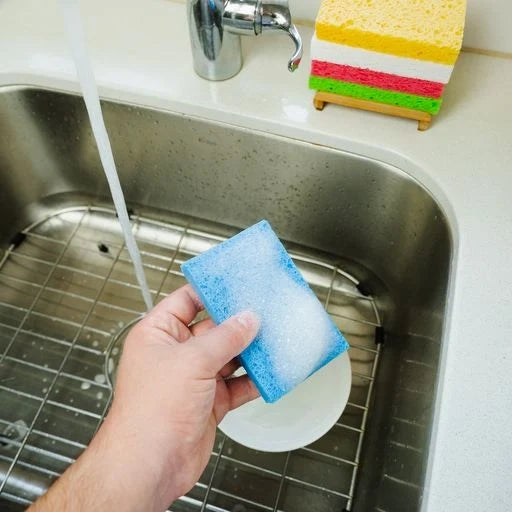 Hand holding a blue sponge under running water in a kitchen sink with colorful sponges on the edge.