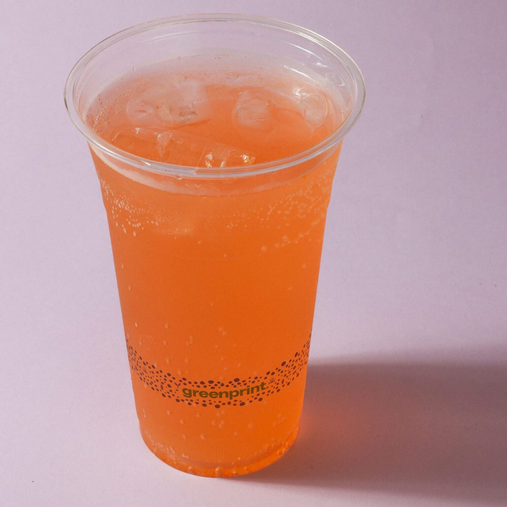 Clear compostable cup with orange beverage and ice on a light purple background