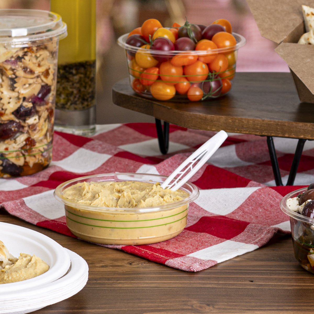 Table with a clear deli bowl of hummus, a deli container of granola, and a clear bowl of cherry tomatoes on a red and white checkered tablecloth.
