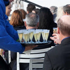 Person serving champagne in compostable champagne glasses from a tray at an outdoor event.