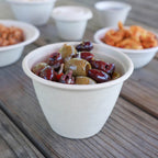 Beige paper bowl filled with olives on a wooden surface with other food items in the background.