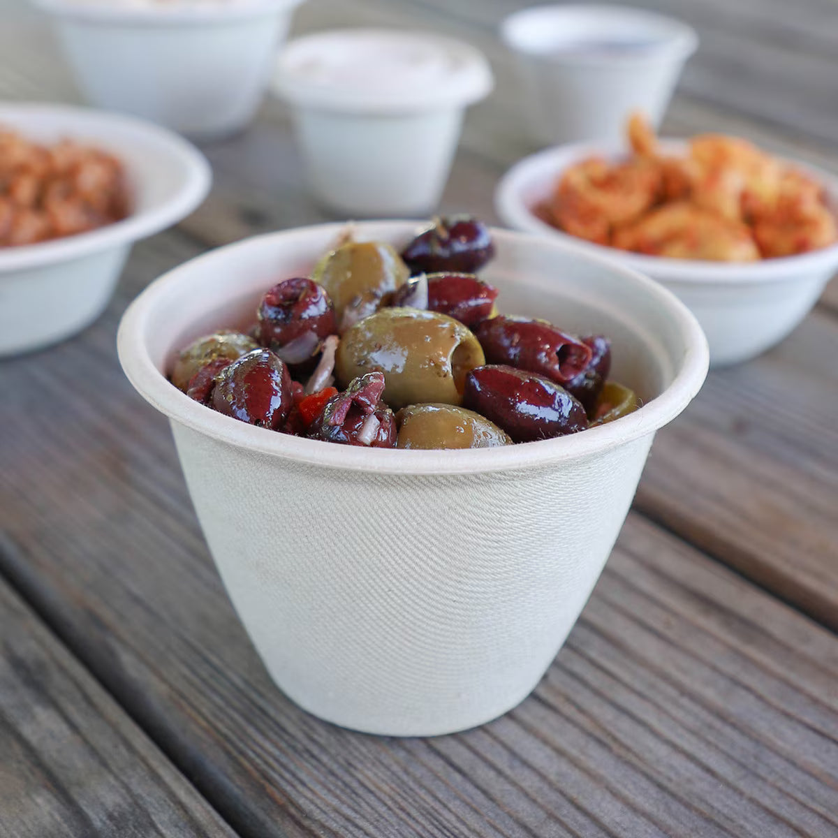 Beige paper bowl filled with olives on a wooden surface with other food items in the background.