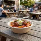 Bowl of pasta with meatballs on a wooden table outdoors, with people and food trucks in the background.