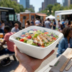 Hand holding a salad container with a food truck and people in the background