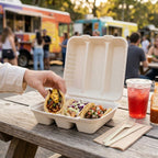 Person picking up a taco from a taco takeout container with food trucks in the background