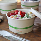 Compostable bowl filled with salad and fruit with a lid on a wooden surface.