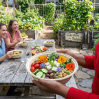 People enjoying a meal outdoors with a 'Garden to Table' sign in the background