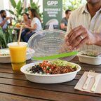 Person opening a World Centric burrito bowl with lid containing a salad, and drink on a wooden table, with 'GPP' branding in the