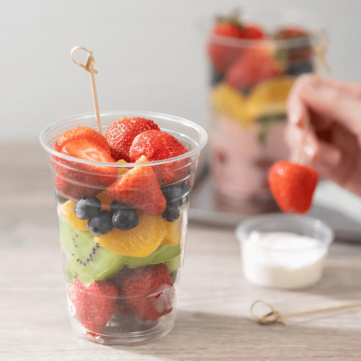 Fruit salad in a clear plastic cup with strawberries, blueberries, and kiwis on a light surface.