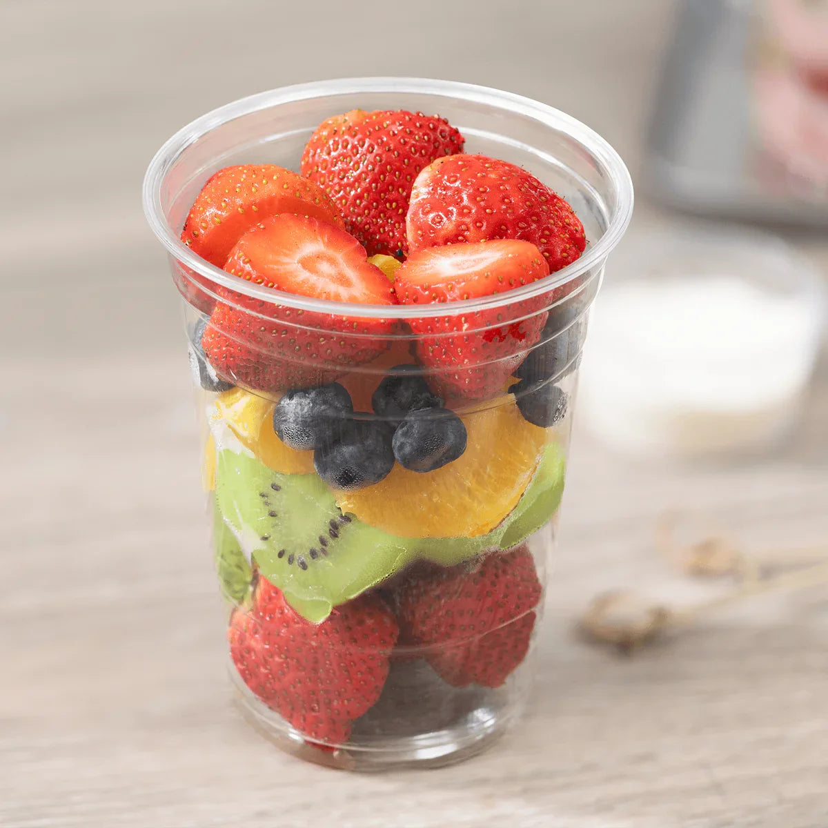 Clear plastic cup filled with sliced strawberries, blueberries, and kiwis on a wooden surface.