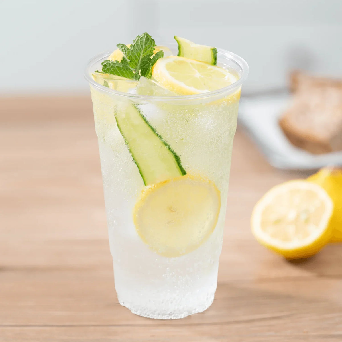 Clear plastic cup of lemon and cucumber infused water on a wooden surface with lemons in the background.