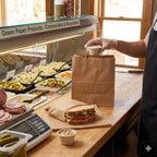 Person preparing a side condiment in a compostable portion cup with lid and placing it into a brown paper bag at a deli counter.