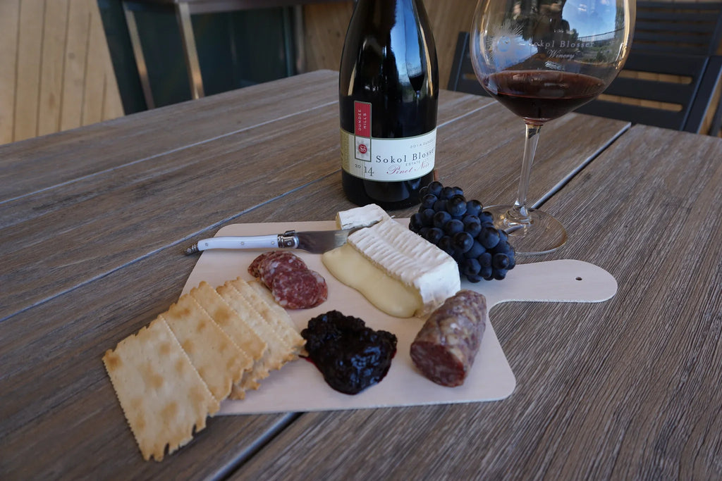 Charcuterie foods on a cheeseboard with a bottle or wine and wine glass on a wooden table.
