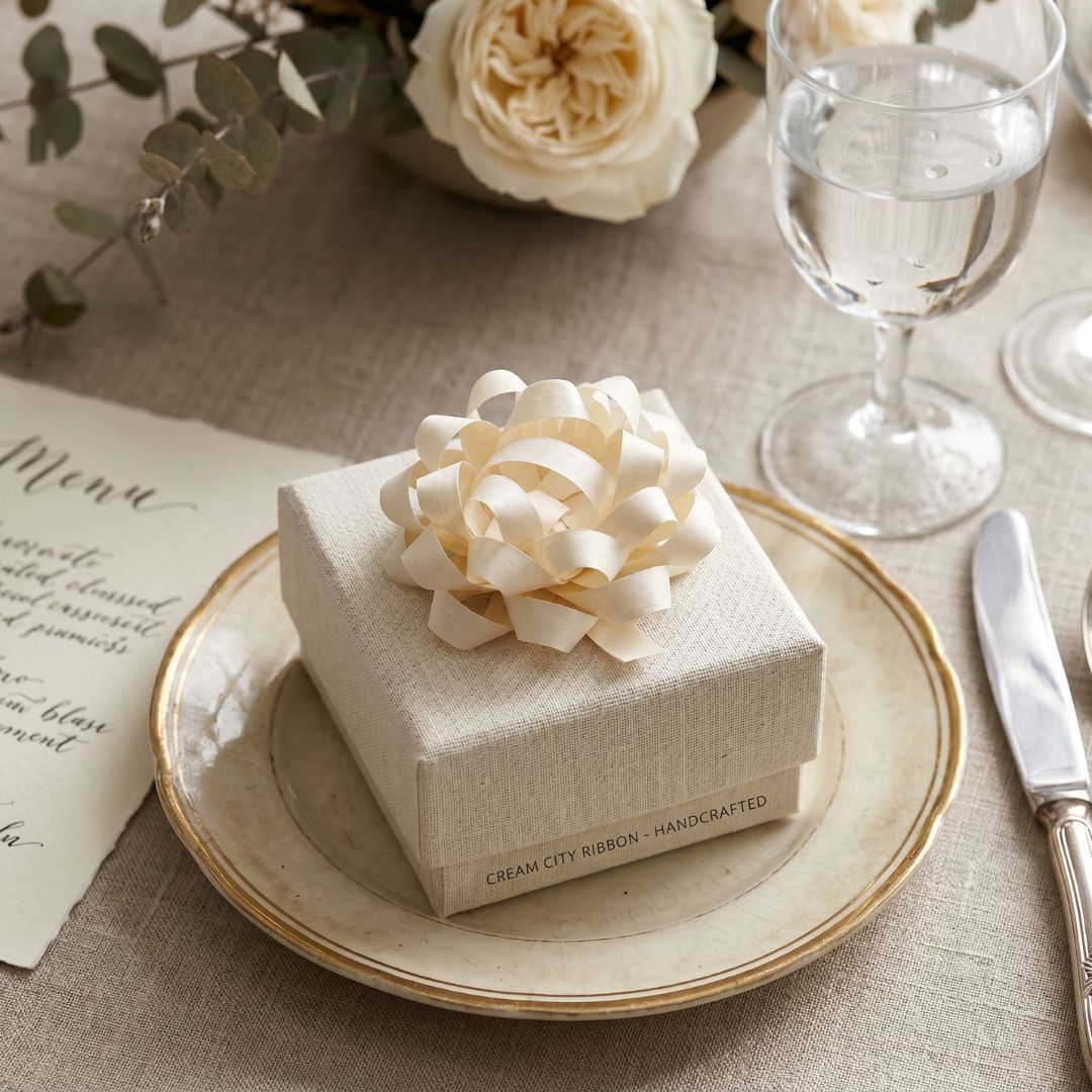 Neatly wrapped gift box with cream ribbon on a table setting with flowers and glasses.