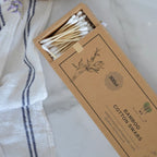 A box of cotton swabs with wooden sticks, shown open with the product visible inside. The box is kraft brown with printed text reading 'BAMBOO COTTON SWABS' and a small logo.