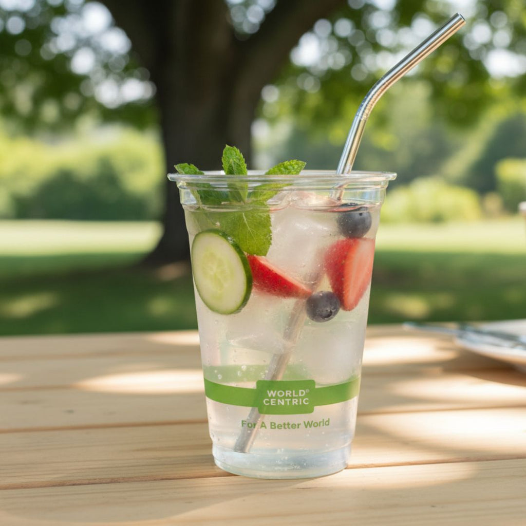 Clear plastic cup with a green label containing a refreshing drink with fruit and a metal straw, set against a blurred natural background.