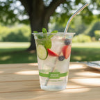 Clear plastic cup with a green label containing a refreshing drink with fruit and a metal straw, set against a blurred natural background.