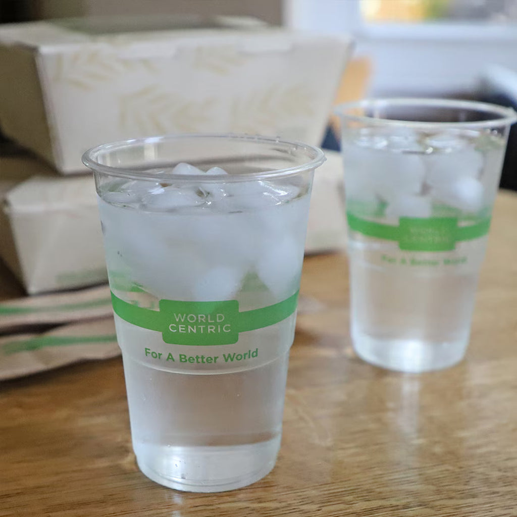 Two clear compostable plastic cups with green labels on a wooden surface.