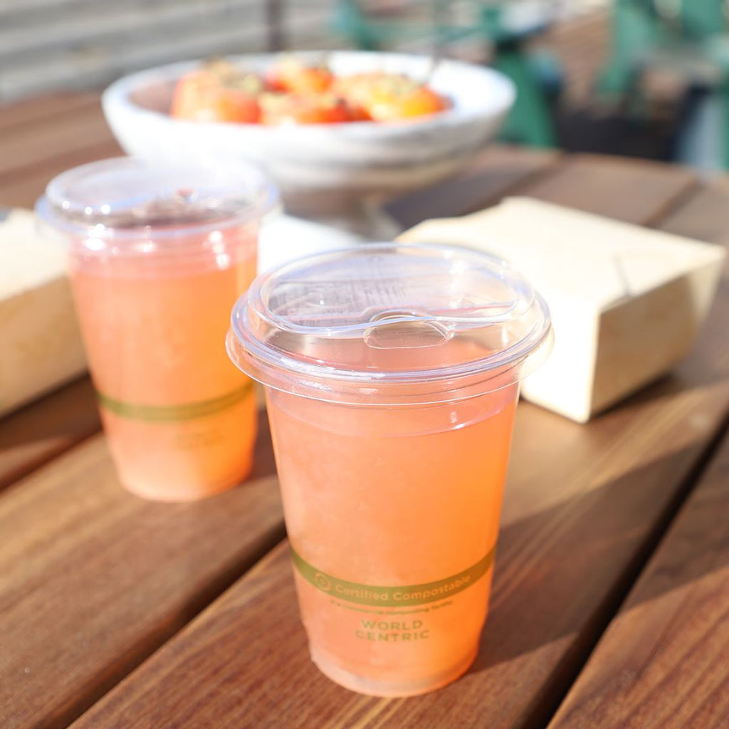 Two compostable cups with lids filled with pink drinks on a wooden table outdoors.
