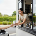 Person receiving a compostable coffee cup from a smiling employee at a drive-thru window.