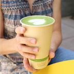 Person holding a compostable coffee cup with green tea inside, wearing a patterned shirt.