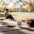 Person sitting at a park table with a World Central Coffee cup and a plate, surrounded by autumn trees.