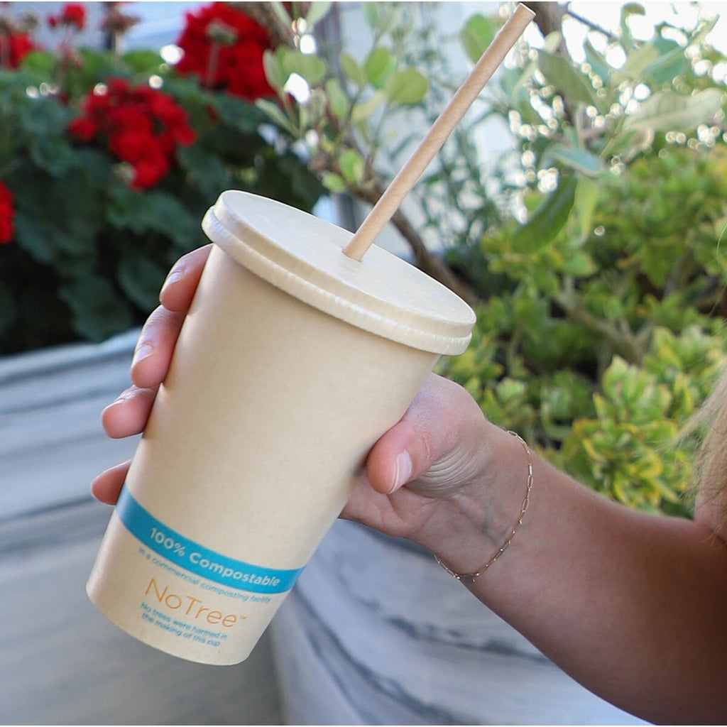 Hand holding a compostable coffee cup with a straw, surrounded by greenery and flowers.