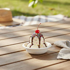 Ice cream sundae with a cherry on top in a white bowl on a wooden table outdoors.