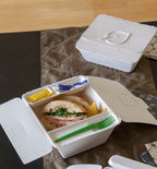 White fiber takeout box that is open containing a sandwich, a green fork, and condiments sitting on a table with a smaller similar takeout box.