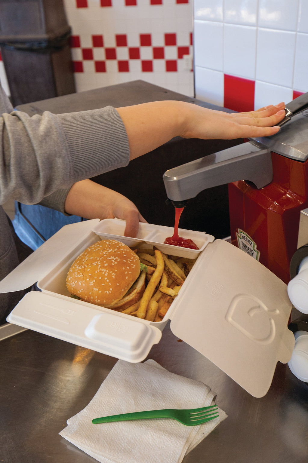 A person filling the sauce compartments of a large takeout box at a condiment station.