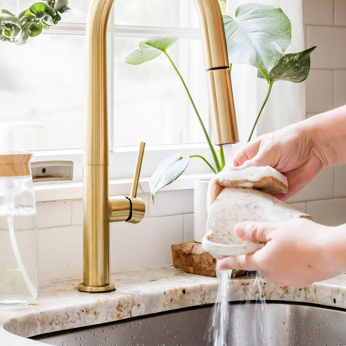 Person washing a stone cup with a scrubber sponge and water from a gold faucet in a kitchen.