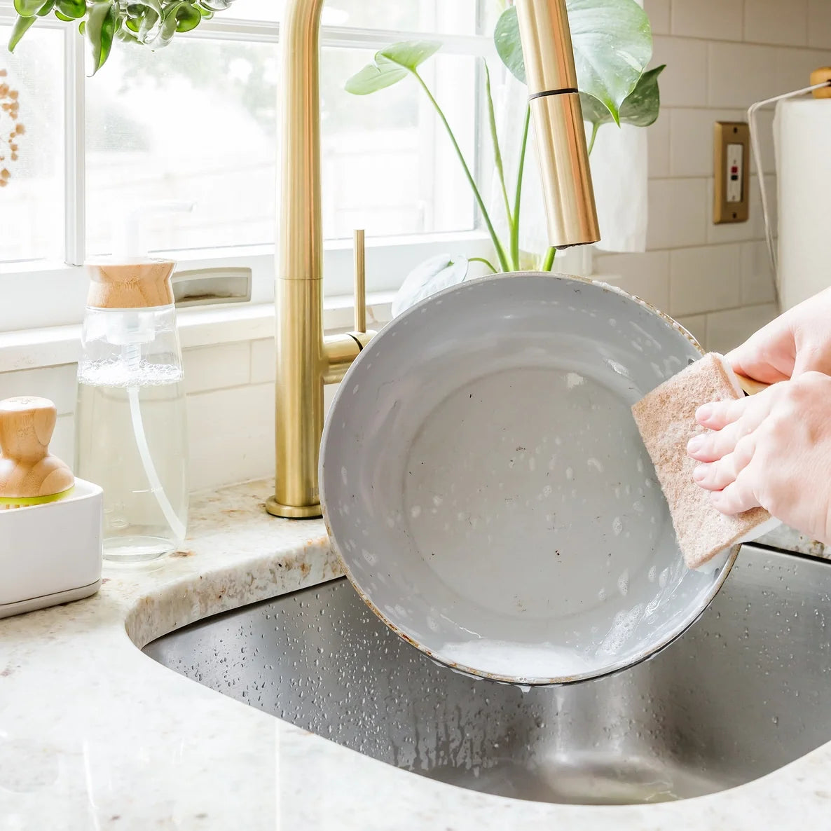 Person washing a gray plate using a dual sided sponge in a kitchen sink with a gold faucet.