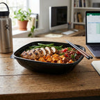 Prepared meal in a plastic black bowl on a wooden desk with a laptop and water bottle in the background.