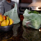 Person holding a green compostable bag next to a bowl of fruit on a kitchen counter.