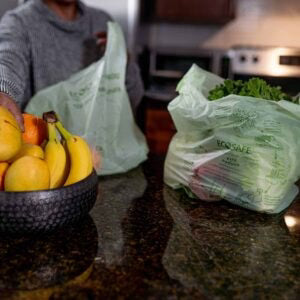 Person holding a green compostable bag next to a bowl of fruit on a kitchen counter.