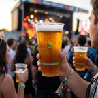 A person at an outdoor concert holds a clear plastic stadium cup of golden beer with a white foam head and a visible Greenware logo with a blurred stage and crowd behind them.