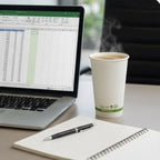 Laptop displaying a spreadsheet on a desk with a coffee cup and notebook.