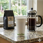 Coffee bag, white cup, and French press on a kitchen counter with a blurred garden view.