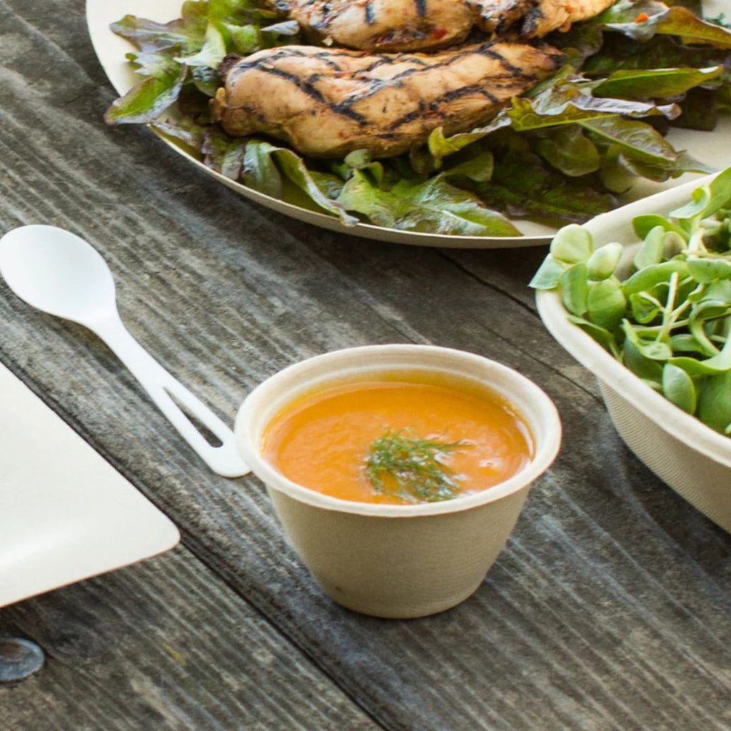 Plated salad with grilled chicken and a small container of soup on a wooden table.