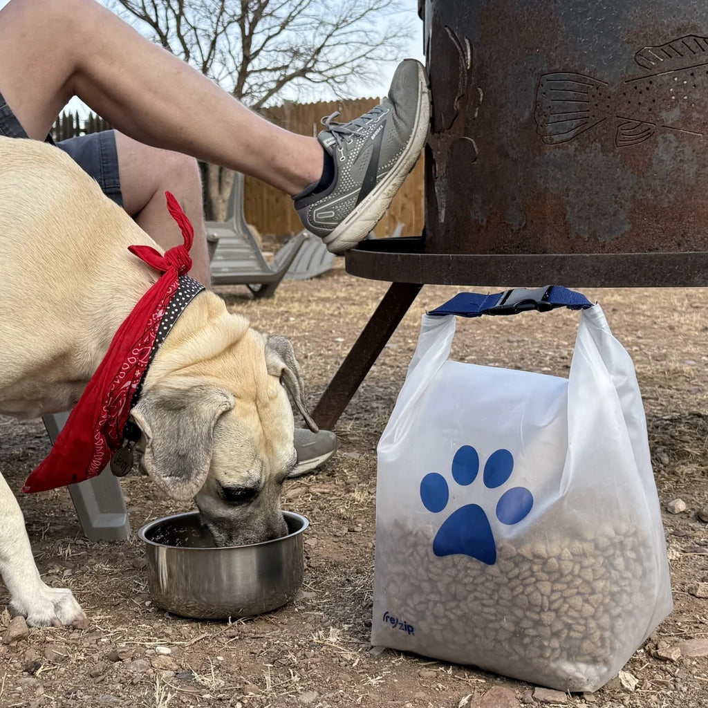 Dog drinking from a bowl next to a bag of dog food with a paw print logo, outdoors.